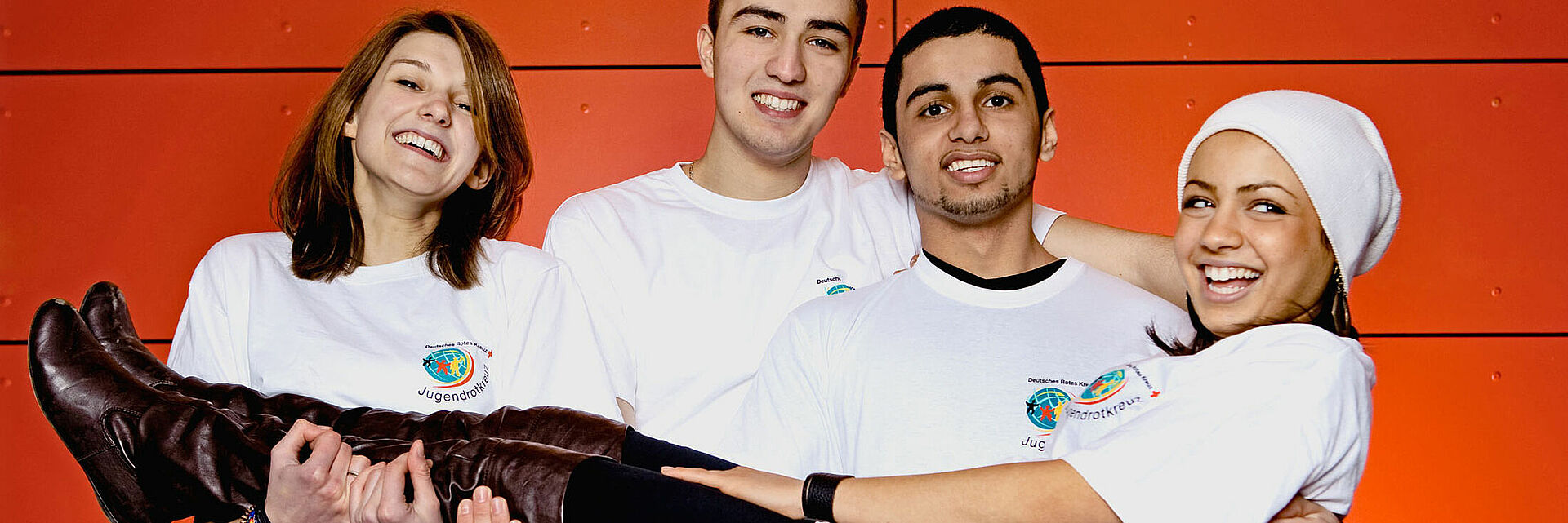 DRK Mittelhessen – Jugendrotkreuz: Engagement mit Herz und Teamgeist Gruppe fröhlicher Jugendlicher des Jugendrotkreuzes in weißen Shirts vor oranger Wand