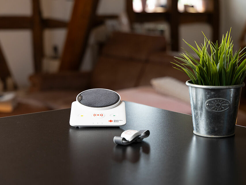 Hausnotrufsystem des DRK Mittelhessen – Sicherheit rund um die Uhr Hausnotrufgerät mit Lautsprecher und Notrufarmband auf einem Tisch