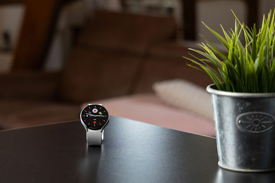 DRK-Mobilruf mit Smartwatch – moderne Sicherheit für unterwegs Moderne DRK-Mobilruf-Smartwatch mit Notruffunktion auf einem Tisch neben einer Pflanze