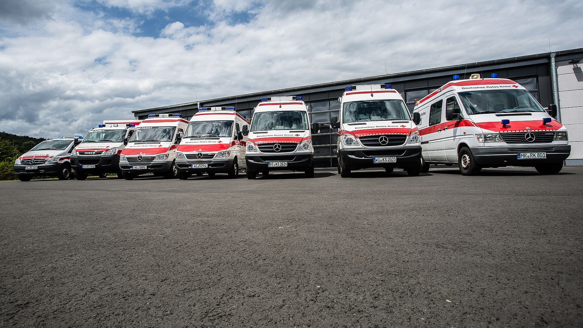 Sanitätszug Marburg-Biedenkopf | DRK-Kreisverband Marburg-Gießen e.V Sieben Einsatzfahrzeuge des DRK-Sanitätszugs Marburg-Biedenkopf stehen aufgereiht vor einer Fahrzeughalle, bereit für den nächsten Einsatz im Katastrophenschutz.