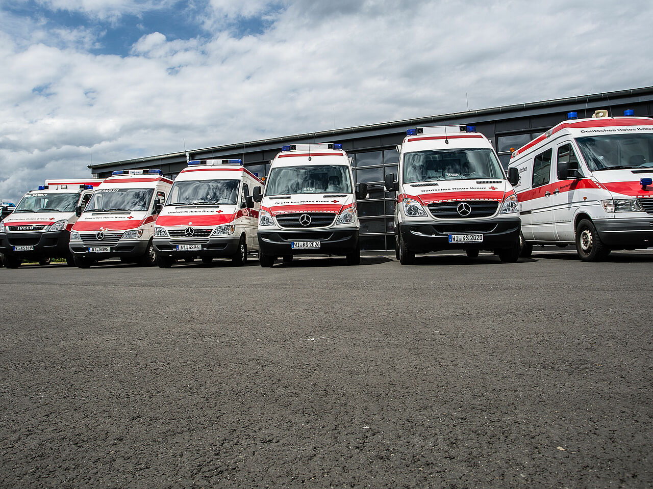 Sanitätszug Marburg-Biedenkopf | DRK-Kreisverband Marburg-Gießen e.V Sieben Einsatzfahrzeuge des DRK-Sanitätszugs Marburg-Biedenkopf stehen aufgereiht vor einer Fahrzeughalle, bereit für den nächsten Einsatz im Katastrophenschutz.