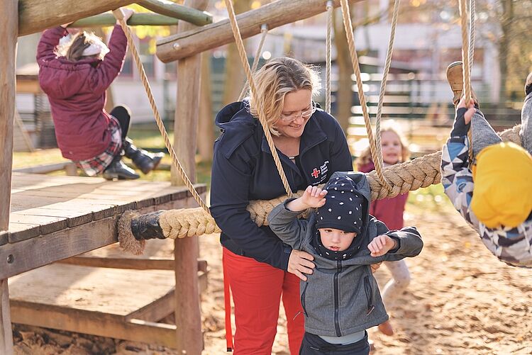 DRK-Kita Mittelhessen – Spielen, Lernen und Vertrauen schaffen Erzieherin unterstützt Kind beim Klettern auf einem Spielplatz der DRK-Kita
