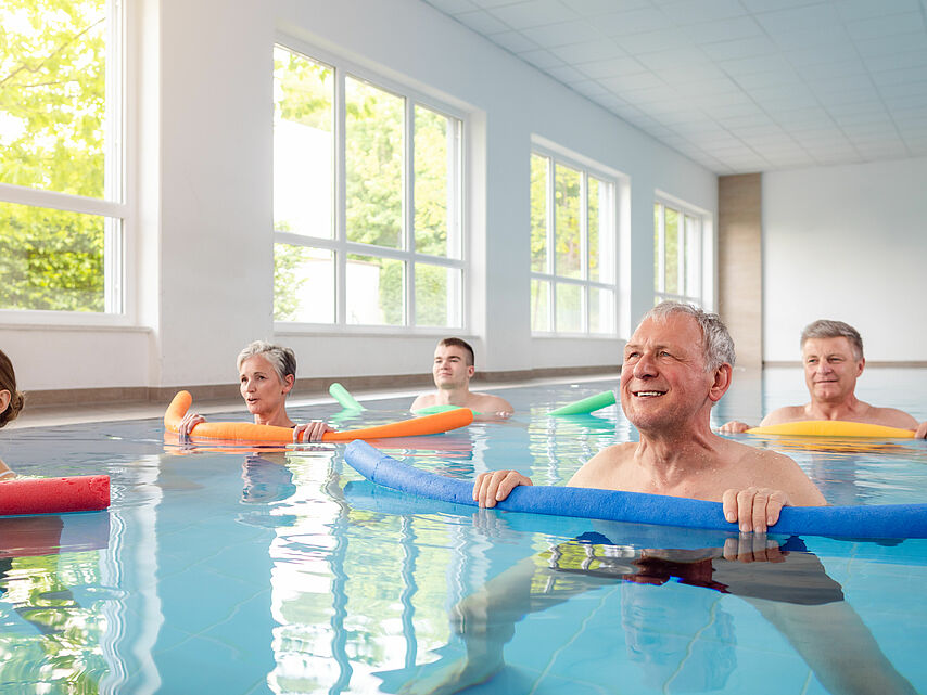 Wassergymnastik beim DRK Mittelhessen – Bewegungstraining im warmen Wasser Gruppe von Menschen macht gemeinsam Wassergymnastik mit Poolnudeln in einem hellen Schwimmbad