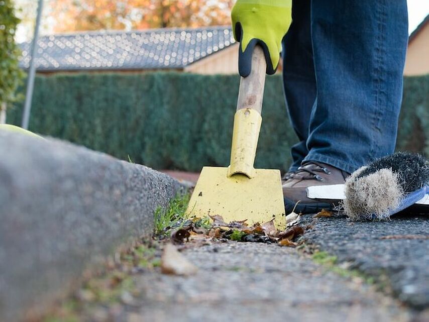 Gehwegreinigung und Außenpflege – DRK Facility Service Mittelhessen Person reinigt mit Schaufel und Besen den Gehweg entlang einer Bordsteinkante