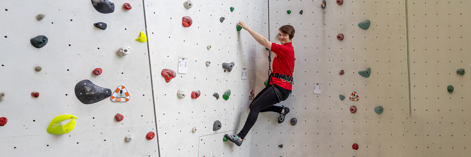 Therapeutisches Klettern beim DRK Mittelhessen Eine Frau in rotem DRK-T-Shirt klettert lächelnd an einer hellen Kletterwand mit bunten Griffen – therapeutisches Klettern beim DRK Mittelhessen.
